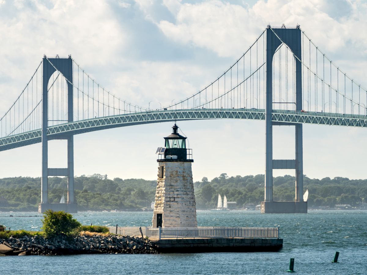 A lighthouse stands on a rocky shore with a tall suspension bridge in the background. Sailboats are visible on the water beneath a partly cloudy sky.