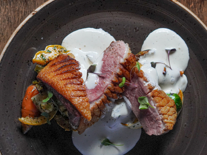 Roasted meat medallions on creamy sauce with herbs and vegetables in a dark bowl.