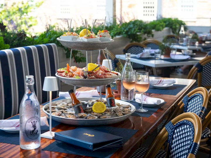 An outdoor restaurant table set with oysters/seafood platters, lemons, bottles, and glasses, with sunlit chairs and umbrellas.