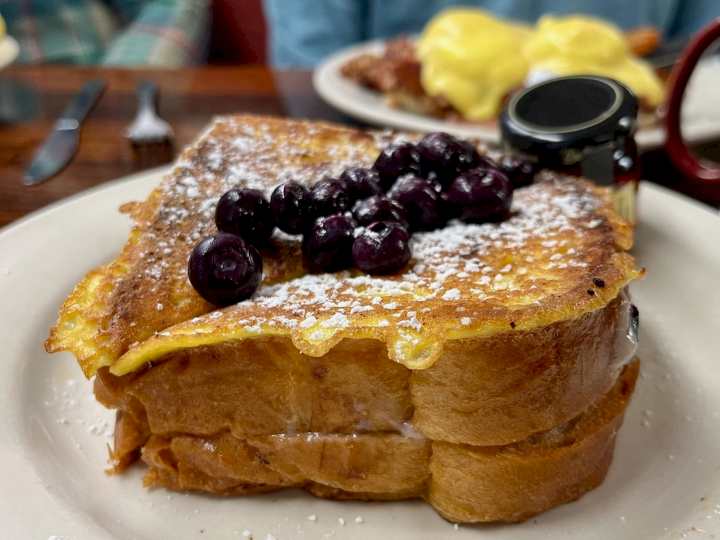 A plate of thick French toast topped with blueberries and powdered sugar, with syrup and a side of eggs in the background.