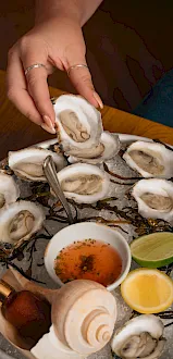 A hand opens fresh oysters on ice, with lemon and dipping sauce nearby.