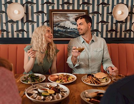 A couple laughs over drinks and food at a cozy restaurant, with nautical dolphin wall decor and wall lamps overhead.