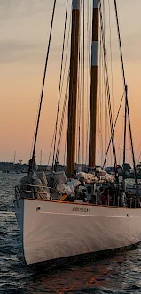 A sailboat named &ldquo;ADIRONDACK IV&rdquo; heads out at sunset, with sails up, crew onboard, and other boats in the harbor.