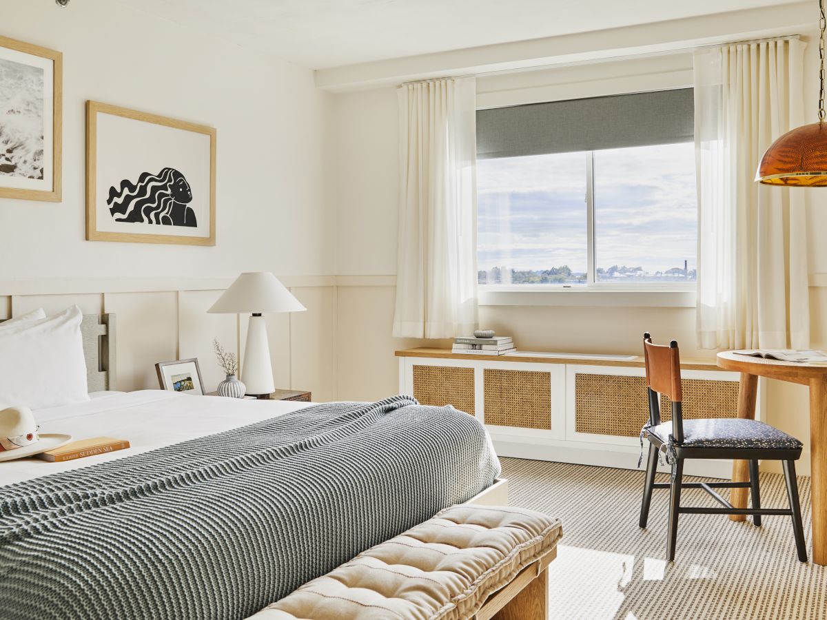 This image shows a modern, well-lit bedroom with a neatly made bed, artwork on the walls, a window with a cityscape view, and a small study table.