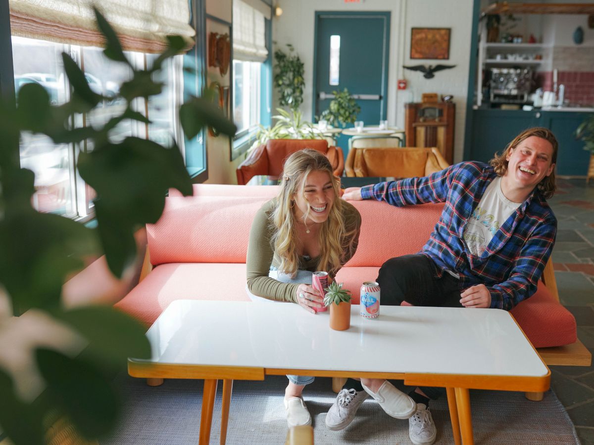 Two people are sitting on a pink couch, laughing and enjoying drinks at a white table in a cozy, well-lit room with plants around.