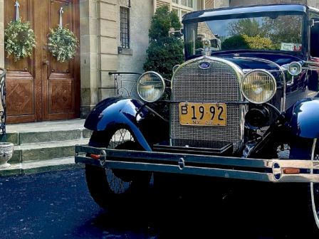 An antique car with the license plate "B 19:92" is parked in front of a building decorated with wreaths on wooden doors.