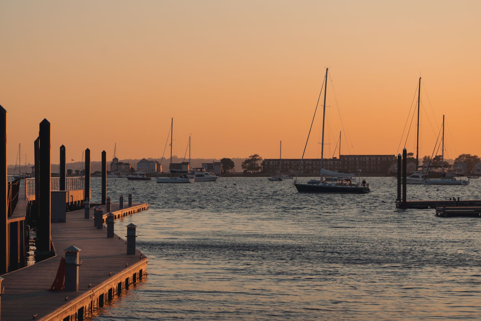 A calm marina at sunset with sailboats moored along a wooden pier, their masts rising over still water and a distant shoreline.