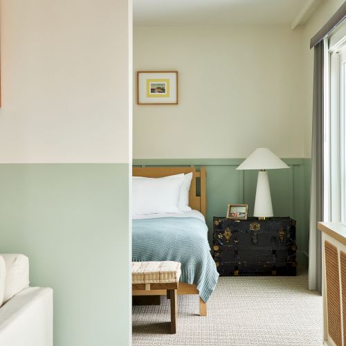 A cozy hotel room with a bed, framed artwork, a dresser, table lamp, and large windows letting in daylight.