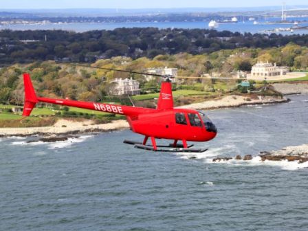 A red helicopter is flying over a coastal area with buildings and vegetation in the background, near the seashore.