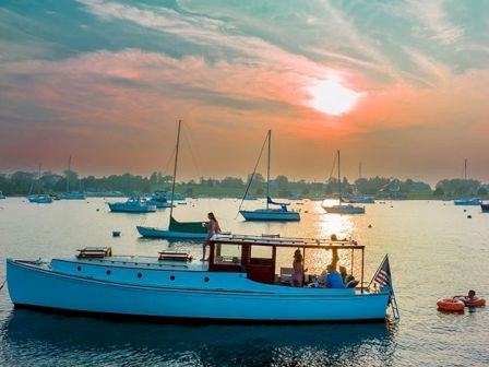 A boat with people on it is floating on the water during a sunset, with other boats in the background and a colorful sky.