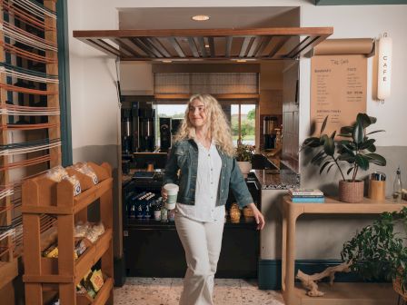 A woman in a caf&eacute; kitchen holds a to-go coffee while espresso machines and a menu board sit behind her.