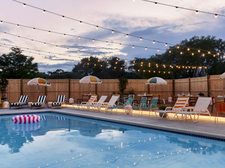 An outdoor pool area with string lights, deck chairs, umbrellas, and inflatables, set against a backdrop of trees and a sunset sky, ending the sentence.