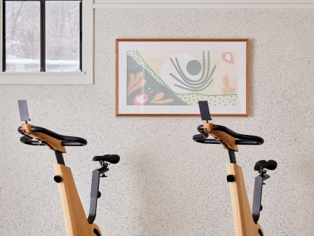 The image shows two stationary exercise bikes in a room with a window and framed wall art.