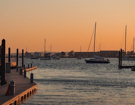 A wooden pier and docked sailboats sit on calm water at sunset, with a quiet riverside skyline in the distance.