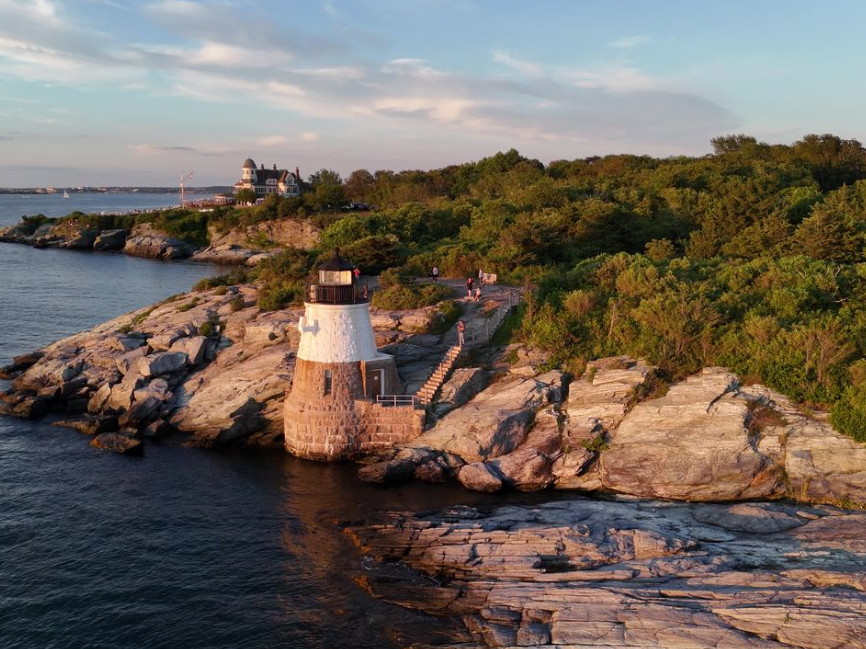A white lighthouse stands on a rocky coastline beside a leafy forest, with small boats sailing on calm blue water.
