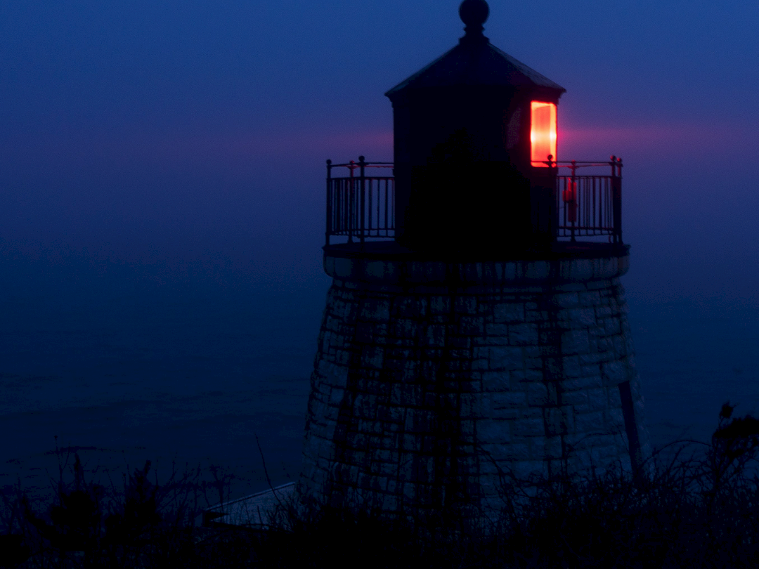 A dark lighthouse on a rocky coast at dusk, its lamp glowing red against the blue foggy sky.