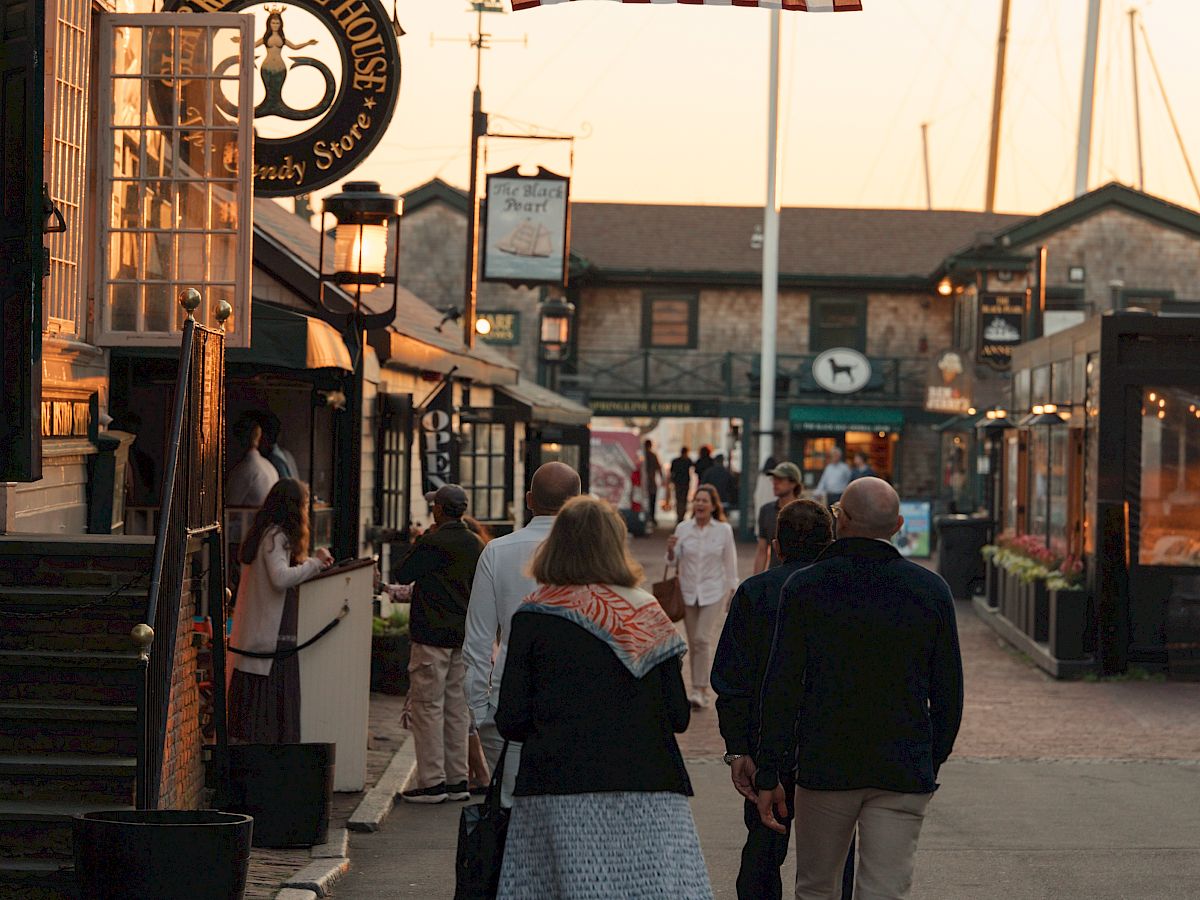 A historic street parade scene with people walking under large American flags on poles at sunset.