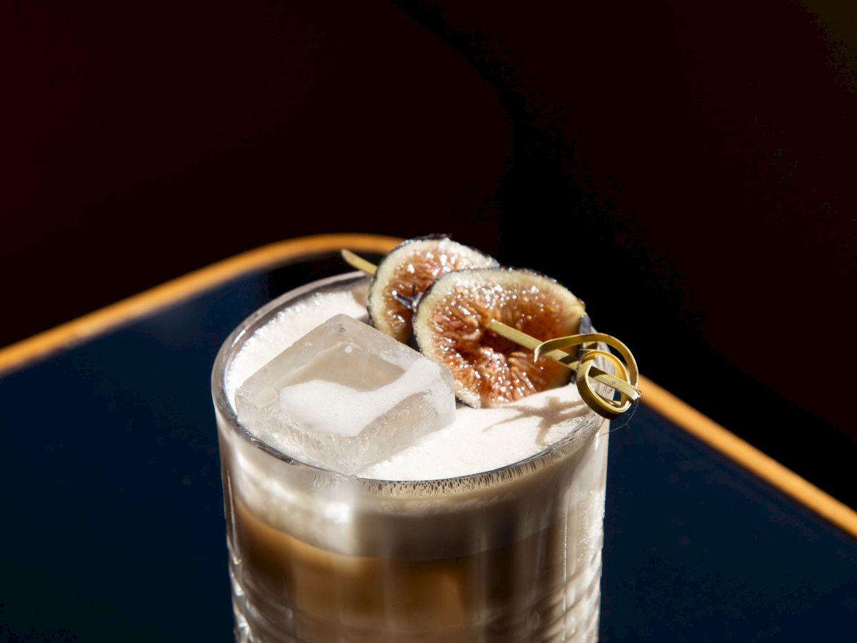 A cocktail in a rocks glass with ice and grilled fig slices, topped with a gold garnish and a drink straw.