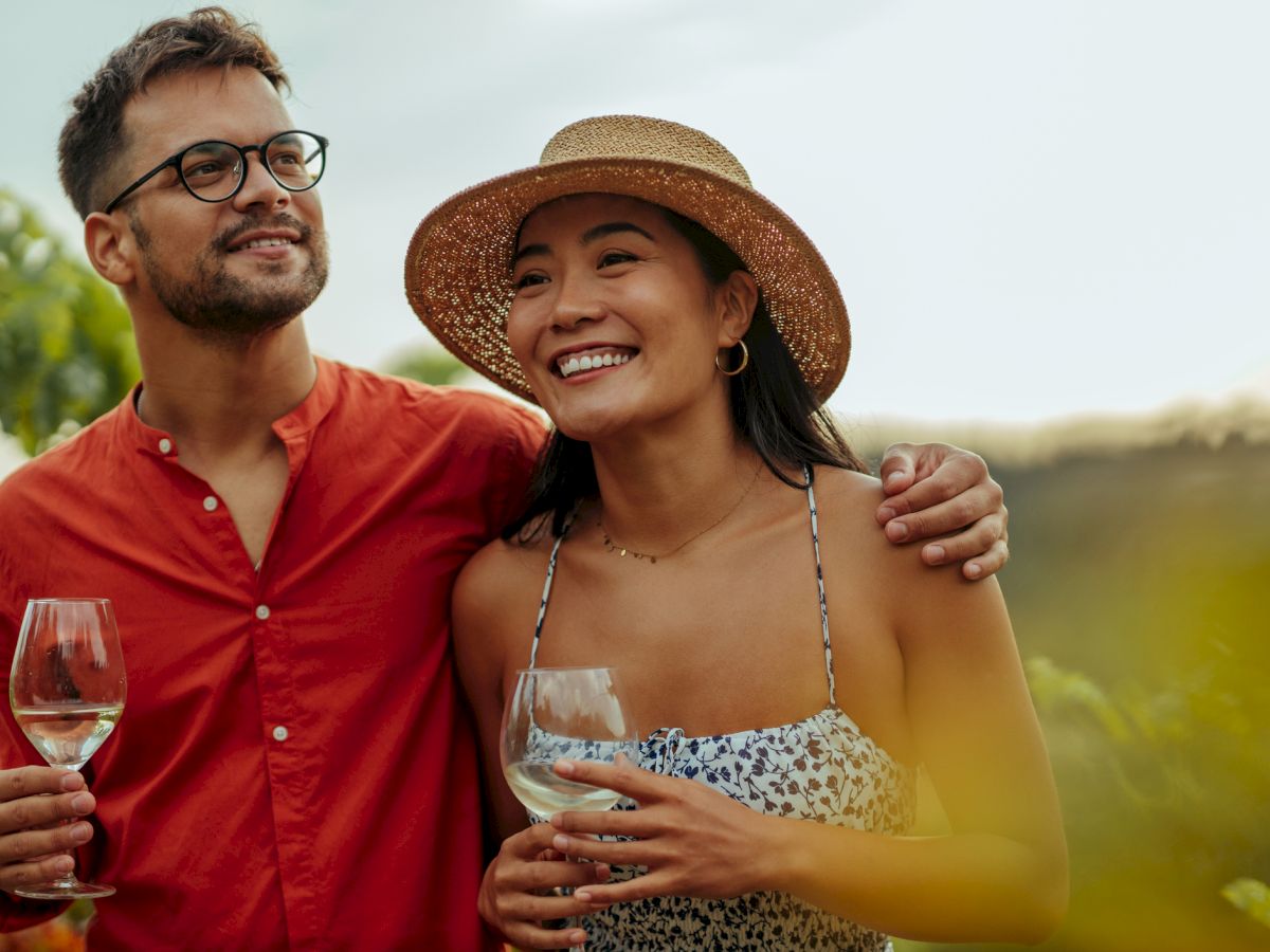 A smiling couple enjoys a glass of wine outdoors, with the man in a red shirt and the woman in a straw hat and patterned dress.