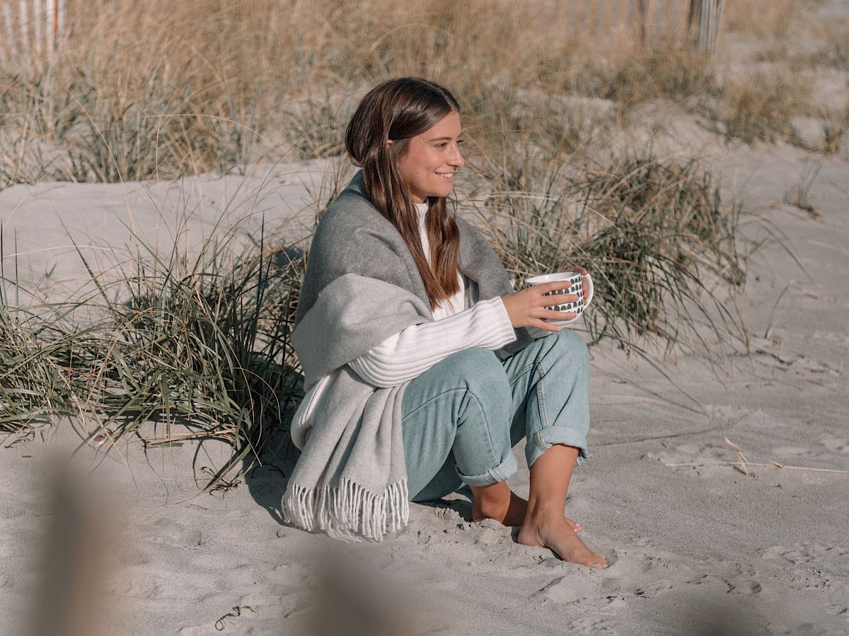 A woman sits on a sandy beach near dune grass, cuddled in a blanket and holding a mug, smiling in the breeze.