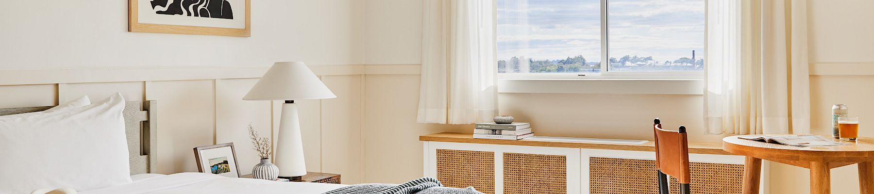 A bright hotel room with a neatly made bed, artwork, a window with a view, and a small table with chairs beneath a warm light fixture.