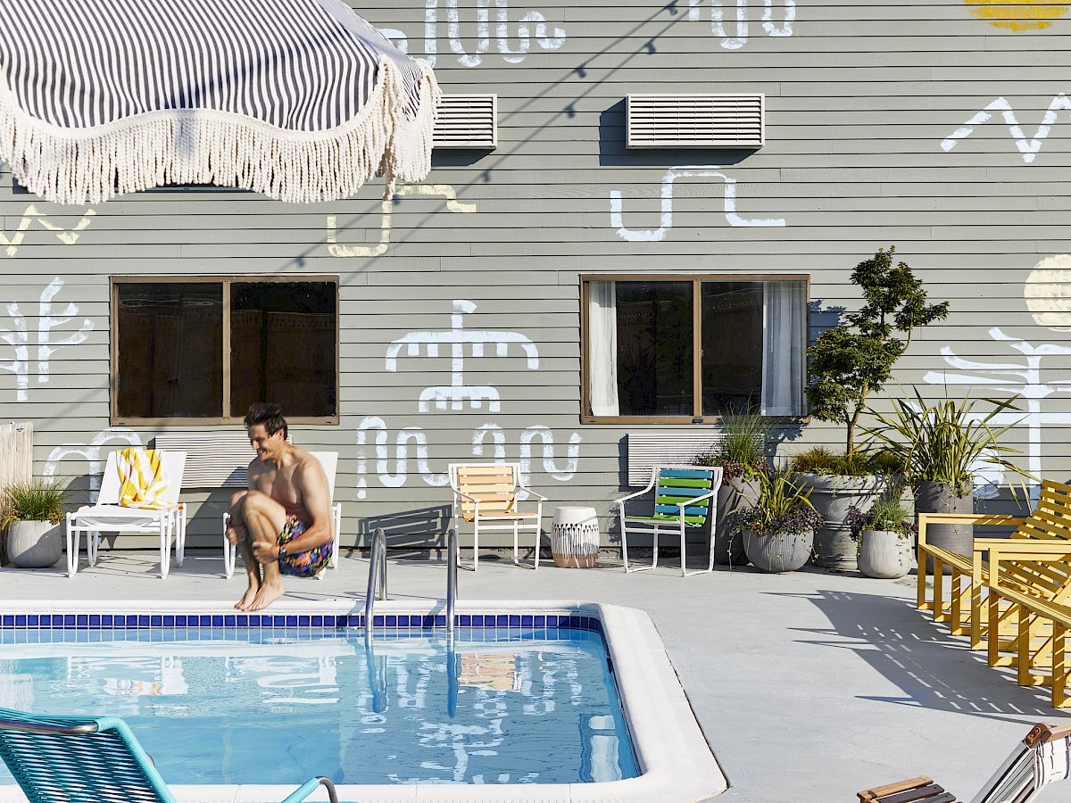 A person jumping into the pool on a sunny day, with a mural of abstract designs on the building wall in the background, chairs, and plants nearby.