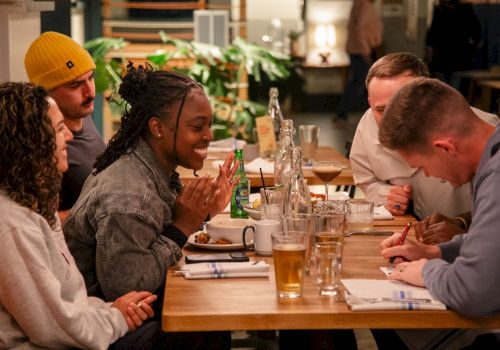 A group of friends cheers at a restaurant table, laughing over drinks and food.