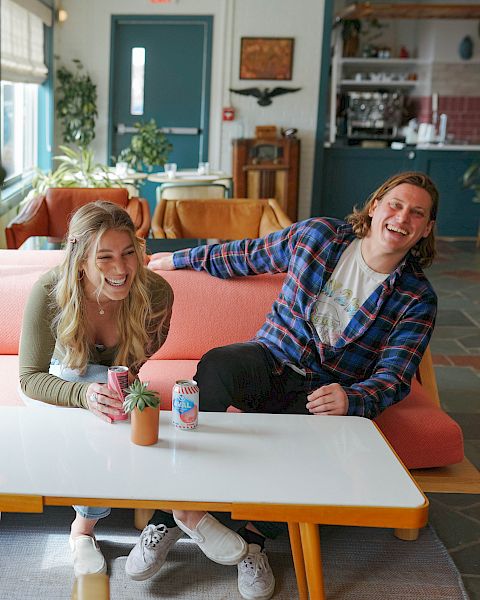 A laughing couple sits on an orange couch in a cozy caf&eacute;, chatting over drinks while plants and decor fill the room.