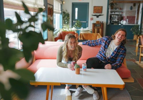 A laughing couple sits on an orange couch in a cozy caf&eacute;, chatting over drinks while plants and decor fill the room.