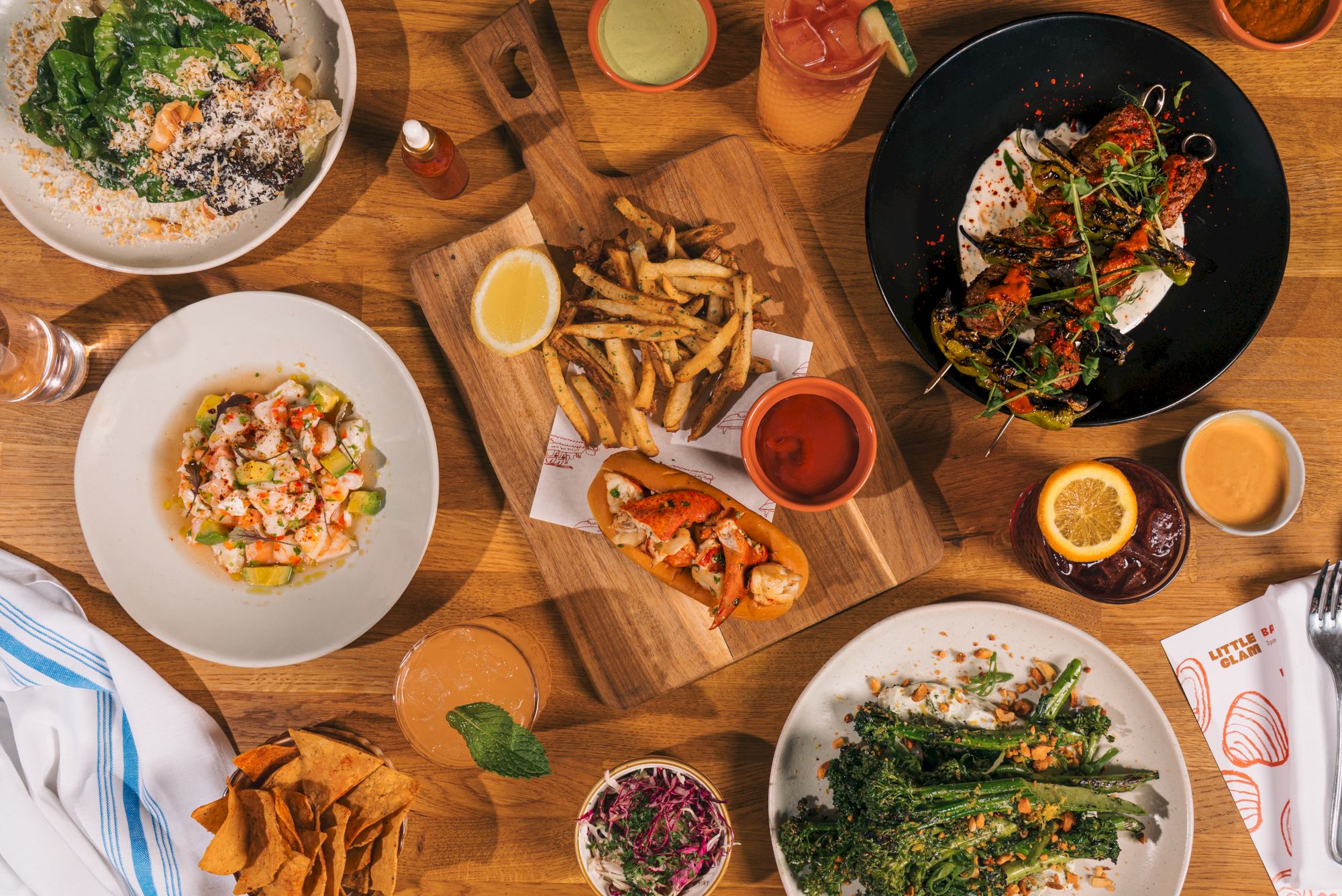 A table spread of tacos/burgers, nacho-style bites, fries, salads, and dips/sauces with lemon and chips, ready to eat.