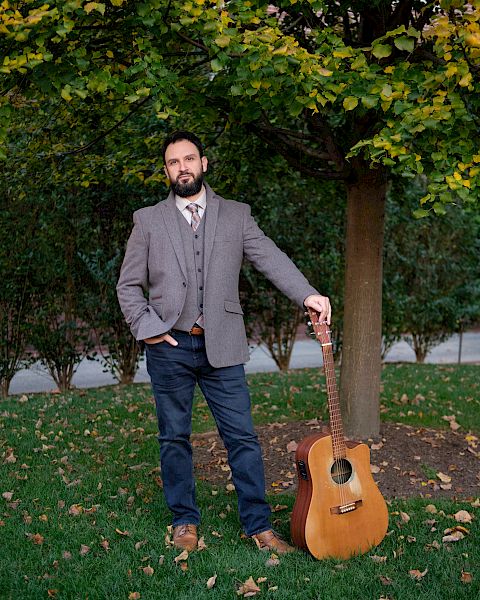 A bearded man in a gray blazer stands outdoors holding an acoustic guitar beside a tree covered in autumn leaves.