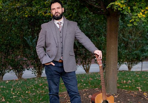 A bearded man in a gray blazer stands outdoors holding an acoustic guitar beside a tree covered in autumn leaves.