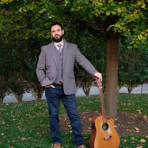 A bearded man in a gray blazer stands outdoors holding an acoustic guitar beside a tree covered in autumn leaves.