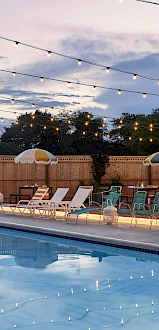 An empty pool area at night with string lights overhead, lounge chairs around the pool, and a few umbrellas along the fence.