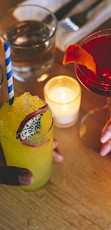 Two hands holding cocktails over a candlelit table. Drinks include a yellow beverage with a dragon fruit slice and a red cocktail.