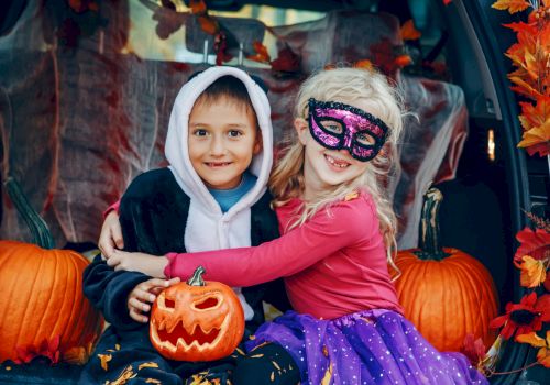 Two children in Halloween costumes, sitting among pumpkins, including a carved jack-o'-lantern, in a decorated setting with autumn leaves.