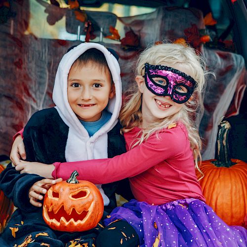 Two children in Halloween costumes, sitting among pumpkins, including a carved jack-o'-lantern, in a decorated setting with autumn leaves.