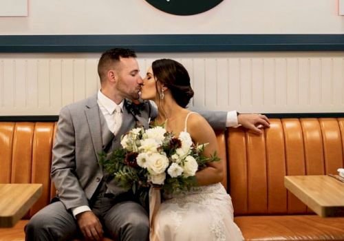 A couple is kissing on a bench in a restaurant, with the bride holding a bouquet. A sign reading 