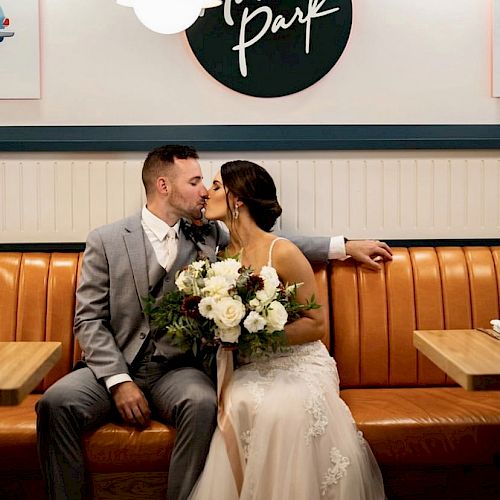 A couple is kissing on a bench in a restaurant, with the bride holding a bouquet. A sign reading 