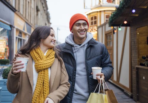 A smiling couple walks through a festive outdoor market. They carry coffee cups and shopping bags, dressed warmly in jackets and scarves.