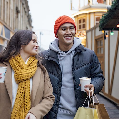 A smiling couple walks through a festive outdoor market. They carry coffee cups and shopping bags, dressed warmly in jackets and scarves.