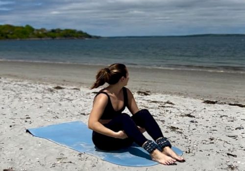 A person sits on a yoga mat on a sandy beach, looking towards the ocean, under a partly cloudy sky.