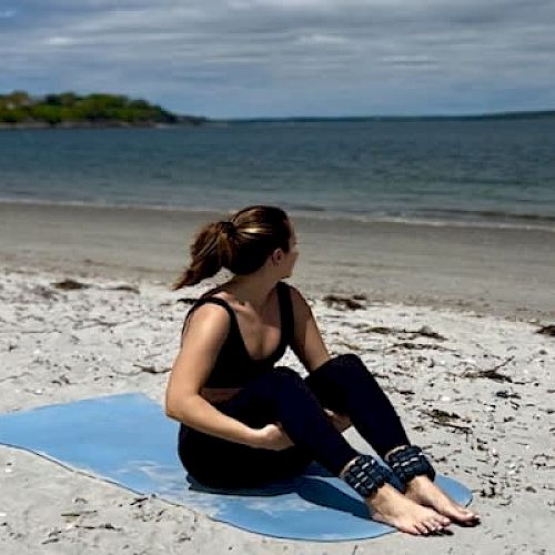 A person sits on a yoga mat on a sandy beach, looking towards the ocean, under a partly cloudy sky.