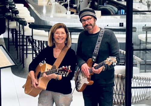 Two people are playing guitars and smiling. They are standing indoors with modern decor in the background.