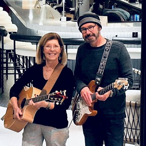 Two people are playing guitars and smiling. They are standing indoors with modern decor in the background.