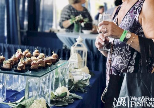 An attendee at the North Fork Food & Wine Festival holds a wine glass, surrounded by appetizers, flowers, and event-related decorations.