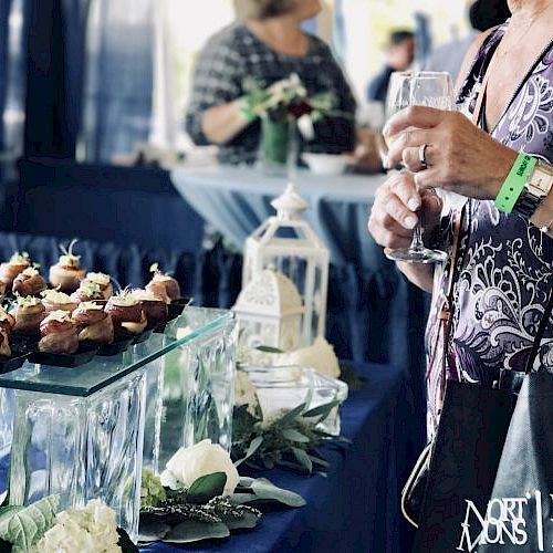 An attendee at the North Fork Food & Wine Festival holds a wine glass, surrounded by appetizers, flowers, and event-related decorations.