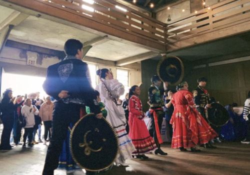 A group of people in traditional attire performing a dance inside a building, with an audience watching them.