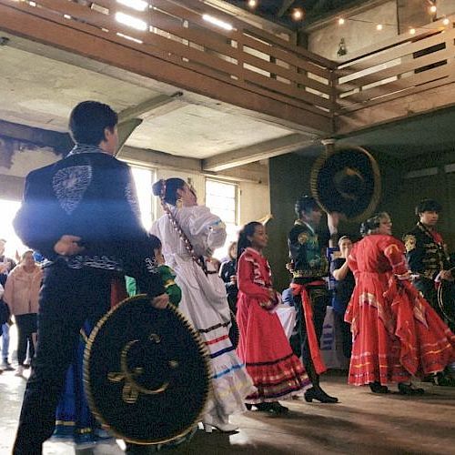 A group of people in traditional attire performing a dance inside a building, with an audience watching them.