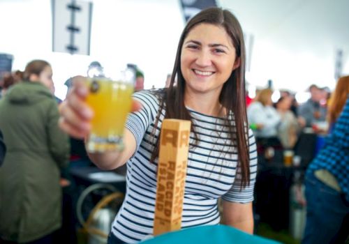 A woman is smiling and holding a glass of beer at an event, with people in the background. She is wearing a striped shirt and standing behind a counter.